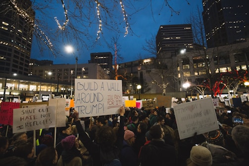 A large group of people gathered in an urban area during the evening, holding various protest signs. The signs feature messages related to political dissent, some specifically targeting policies associated with Donald Trump. Buildings and trees decorated with lights form the backdrop, and the setting appears to be in a city square.