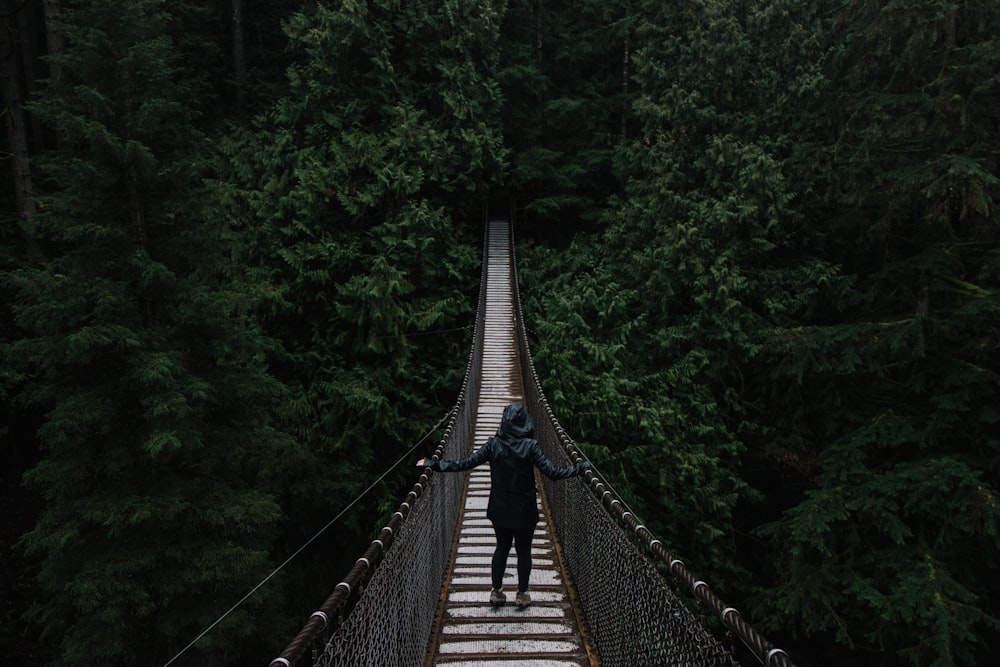 person in black hoodie on wooden bridge surrounded by green leafed trees