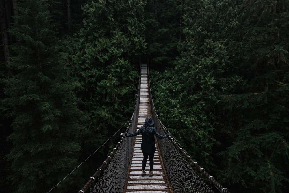 pessoa em capuz preto na ponte de madeira cercada por árvores de folhas verdes
