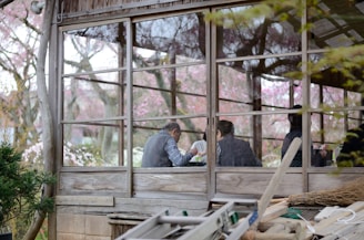 A rustic wooden structure with large windows reveals several people sitting inside, engaged in conversation or activity. The background showcases a lush exterior with trees and pink blossoms, suggesting a serene, natural setting.