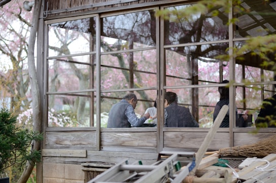 A rustic wooden structure with large windows reveals several people sitting inside, engaged in conversation or activity. The background showcases a lush exterior with trees and pink blossoms, suggesting a serene, natural setting.
