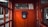 An old-fashioned black rotary dial telephone is mounted on the vibrant red interior wall of a vintage phone booth. The booth has glass panel windows, providing a glimpse of an outdoor setting with trees, a wooden building, and a paved road.