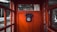 An old-fashioned black rotary dial telephone is mounted on the vibrant red interior wall of a vintage phone booth. The booth has glass panel windows, providing a glimpse of an outdoor setting with trees, a wooden building, and a paved road.