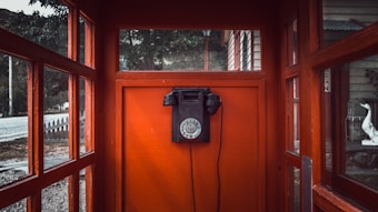 An old-fashioned black rotary dial telephone is mounted on the vibrant red interior wall of a vintage phone booth. The booth has glass panel windows, providing a glimpse of an outdoor setting with trees, a wooden building, and a paved road.
