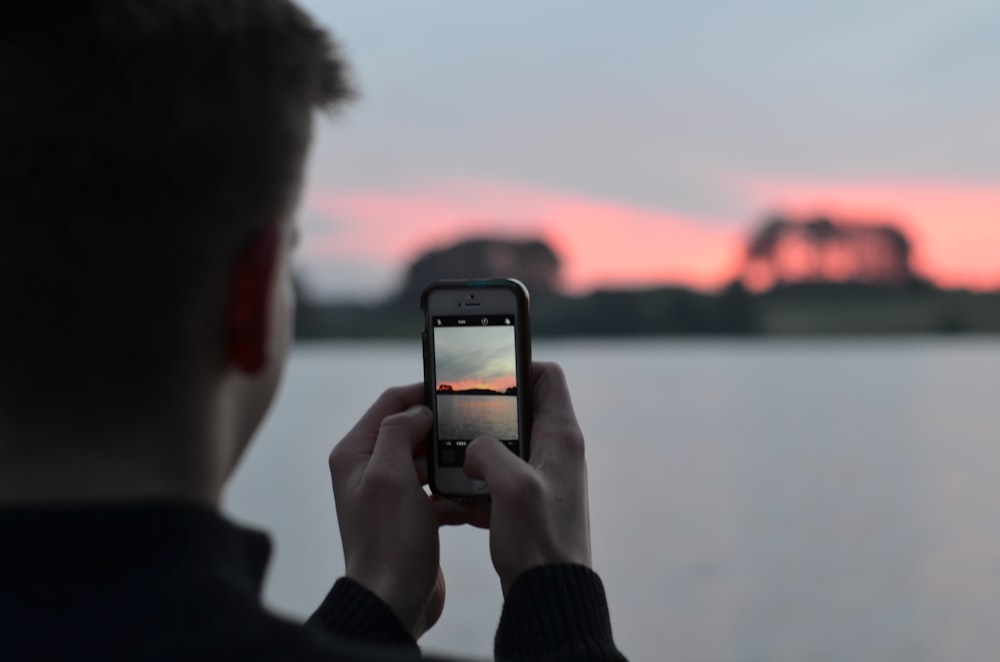 selective focus photography of person using smartphone camera taking photo of blue body of water