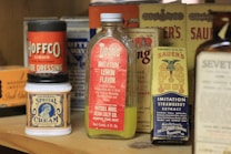 A variety of antique food and household product containers are arranged on a wooden shelf. The items include a bottle labeled Dean's Imitation Lemon Flavor, a jar of Universal Special Cream, and a box of Sauers Imitation Strawberry Extract. There are also containers for other products such as Hoffco Liquid Shoe Dressing and various tins and boxes in the background.