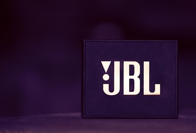 A box-shaped object with the letters JBL prominently displayed in bold, stylish font. The background is blurred and predominantly dark purple, creating a sleek, modern aesthetic.