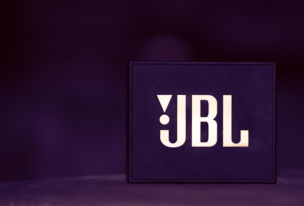 A box-shaped object with the letters JBL prominently displayed in bold, stylish font. The background is blurred and predominantly dark purple, creating a sleek, modern aesthetic.