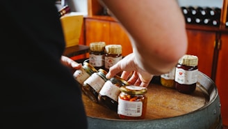 Several jars of preserves are placed on a round wooden surface. A person is reaching into these jars with their right hand. The jars have different types of contents, and the background consists of wooden cabinets and a shelf with bottles.