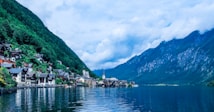A picturesque lakeside village nestled at the foot of lush, green mountains. The water reflects the vibrant landscape and sky above, creating a mirror-like surface. Traditional alpine houses with characteristic architecture are clustered along the shoreline. The sky is partially cloudy, adding texture to the scenery.