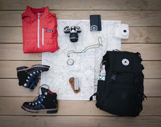A selection of outdoor and travel items neatly arranged on a wooden surface, including a red jacket, two pairs of black hiking boots, a black backpack, a map, three cameras, a metal water bottle, a compass, a wristwatch, and a folding knife.