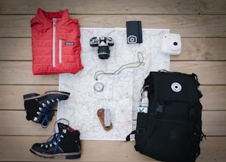 black hiking backpack near white Fujifilm instax mini camera near black leather boots, red half-zip jacket, gray pocket watch on white map