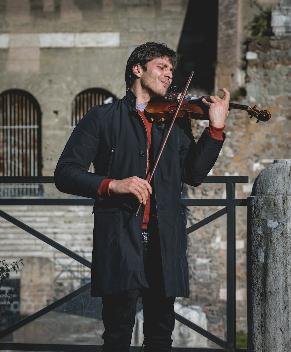 A man in a black coat playing the violet with his eyes closed in front of an old building