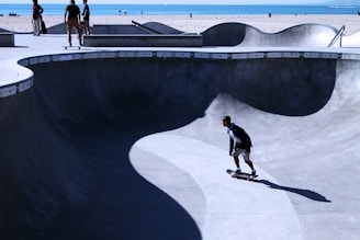 A smooth concrete skate bowl with skaters enjoying the flow under a bright blue sky.
