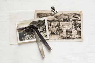 A vintage postcard and a black-and-white photograph are placed on top of each other. A binder clip holds them together, and a ribbon is draped across the photo. The postcard features an image of a patio with tables and chairs under striped umbrellas, and the photograph shows a rocky landscape.