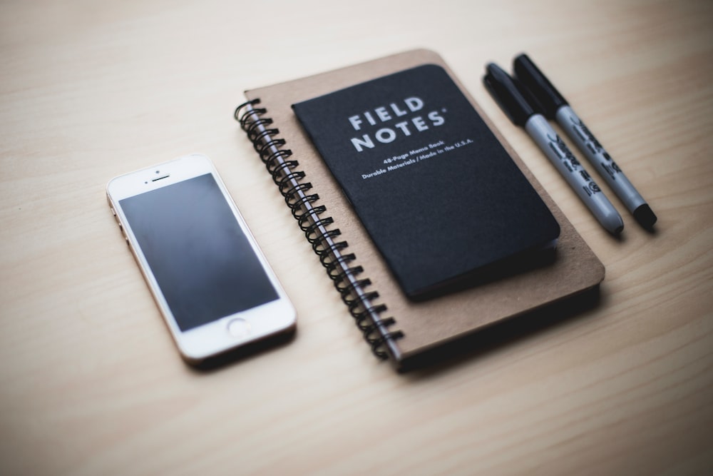 A Field Notes memo book on top of a larger spiral notebook with two sharpies on the right and a phone on the left