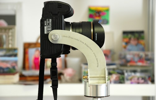 A close-up view of a DSLR camera mounted on a specialized metal bracket. The lens is prominently seen, labeled with 'ASPHERICAL.' The background features several blurred photographs and frames, suggesting a living space or studio environment.
