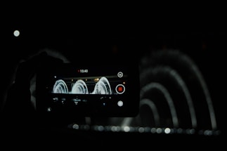 A dark scene featuring a smartphone capturing a video of an illuminated water fountain creating arcs with its jets. The phone's screen prominently displays the recording interface with elapsed time, a red recording button, and additional control icons. Water spray pattern is visible beyond the phone screen, emphasizing the elegance of the fountain display.