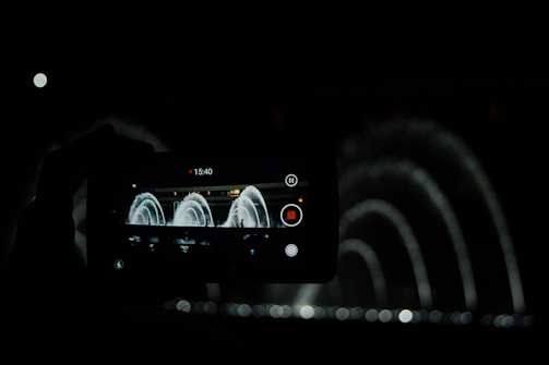 A dark scene featuring a smartphone capturing a video of an illuminated water fountain creating arcs with its jets. The phone's screen prominently displays the recording interface with elapsed time, a red recording button, and additional control icons. Water spray pattern is visible beyond the phone screen, emphasizing the elegance of the fountain display.
