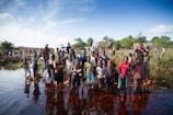 A large group of children and adults stand near a body of water. Some children are wading in the water with bricks beneath their feet. The background includes more people and simple huts with a flag visible. The sky is clear with some clouds, and the surrounding area is lush with greenery.