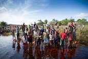A large group of children and adults stand near a body of water. Some children are wading in the water with bricks beneath their feet. The background includes more people and simple huts with a flag visible. The sky is clear with some clouds, and the surrounding area is lush with greenery.