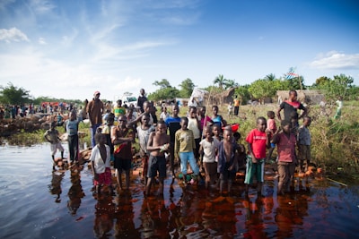 A large group of children and adults stand near a body of water. Some children are wading in the water with bricks beneath their feet. The background includes more people and simple huts with a flag visible. The sky is clear with some clouds, and the surrounding area is lush with greenery.