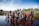 A large group of children and adults stand near a body of water. Some children are wading in the water with bricks beneath their feet. The background includes more people and simple huts with a flag visible. The sky is clear with some clouds, and the surrounding area is lush with greenery.