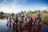A large group of children and adults stand near a body of water. Some children are wading in the water with bricks beneath their feet. The background includes more people and simple huts with a flag visible. The sky is clear with some clouds, and the surrounding area is lush with greenery.
