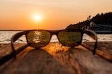 A pair of sunglasses is placed on a wooden surface, with a view of a serene lake during sunset. The sun casts a warm glow over the water, and a boat is visible on the right side near a line of trees. The image is focused on the sunglasses, creating a blurred background that emphasizes the golden hues reflected in the lenses.