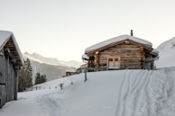 A wooden cabin sits amid snow-covered surroundings, with snowy mountains in the background. The cabin has a chimney and is surrounded by deep snow. Tracks and footprints are visible in the snow, indicating recent activity. Another wooden structure is partially visible to the left.
