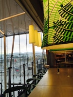 Inside a modern cafe or restaurant with large windows overlooking a busy urban area. Green-patterned overhead lighting complements wooden tables and chairs, creating a cozy yet vibrant ambiance. The outside view shows a populated street with vehicles and buildings against a backdrop of mountains.