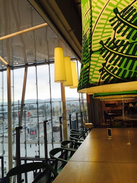 Inside a modern cafe or restaurant with large windows overlooking a busy urban area. Green-patterned overhead lighting complements wooden tables and chairs, creating a cozy yet vibrant ambiance. The outside view shows a populated street with vehicles and buildings against a backdrop of mountains.