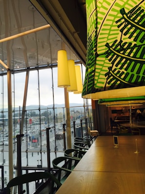 Inside a modern cafe or restaurant with large windows overlooking a busy urban area. Green-patterned overhead lighting complements wooden tables and chairs, creating a cozy yet vibrant ambiance. The outside view shows a populated street with vehicles and buildings against a backdrop of mountains.