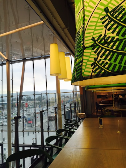 Inside a modern cafe or restaurant with large windows overlooking a busy urban area. Green-patterned overhead lighting complements wooden tables and chairs, creating a cozy yet vibrant ambiance. The outside view shows a populated street with vehicles and buildings against a backdrop of mountains.