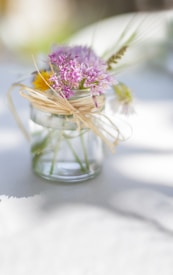 A small glass jar filled with a bouquet of wildflowers, tied with a piece of twine. The flowers include purple and yellow blooms, with a soft green background giving a blurred effect. Light and shadow play delicately on a white surface, creating a peaceful and simple arrangement.