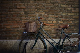 A classic bicycle with a wicker basket attached to the front is parked against a rustic brick wall. The bicycle has a dark green frame and brown leather seat and handlebars, adding to its vintage appeal.