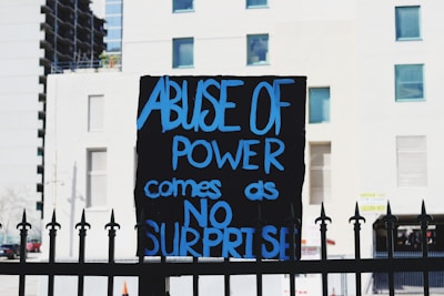 A sign with the message 'Abuse of power comes as no surprise' written in bold blue letters against a dark background. The sign is mounted on a wrought iron fence in front of a white building with several windows.