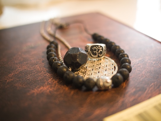 A set of black beads, a silver ring with an engraved symbol, and a decorative metallic disc are arranged on a textured surface. The beads form a necklace or bracelet, and the ring features an intricate motif. The metallic disc beneath the ring has a geometric pattern.