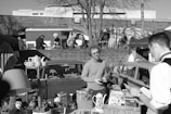 A bustling outdoor market scene features a variety of vintage items for sale, including a collection of kitchenware, books, and potted plants. Shoppers and vendors are actively engaged in conversation next to a car parked near a small table. The background includes a brick wall with people walking and exploring stalls against an urban cityscape.