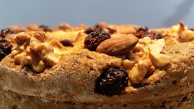 A close-up of a baked good topped with various nuts and dried fruits, including walnuts, almonds, and raisins. The surface appears golden-brown and slightly textured, suggesting a crisp exterior.