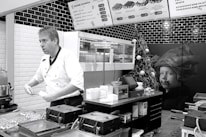 A chef in a white uniform is standing in a modern kitchen or food stall, possibly preparing food. Wall-mounted menu boards display food items and prices. The decor includes black and white tiles, and there is a Christmas tree with ornaments as well as a large picture of a child on the wall.