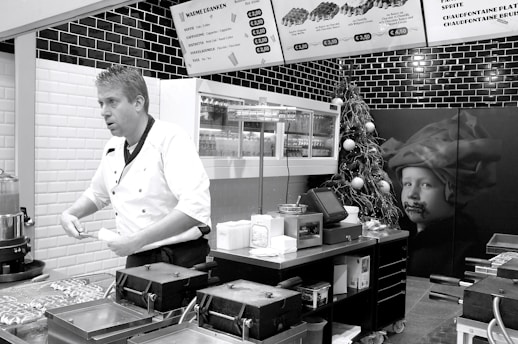 A chef in a white uniform is standing in a modern kitchen or food stall, possibly preparing food. Wall-mounted menu boards display food items and prices. The decor includes black and white tiles, and there is a Christmas tree with ornaments as well as a large picture of a child on the wall.