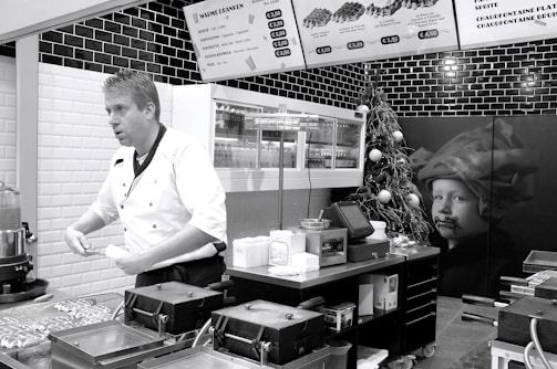 A chef in a white uniform is standing in a modern kitchen or food stall, possibly preparing food. Wall-mounted menu boards display food items and prices. The decor includes black and white tiles, and there is a Christmas tree with ornaments as well as a large picture of a child on the wall.