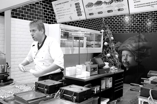 A chef in a white uniform is standing in a modern kitchen or food stall, possibly preparing food. Wall-mounted menu boards display food items and prices. The decor includes black and white tiles, and there is a Christmas tree with ornaments as well as a large picture of a child on the wall.