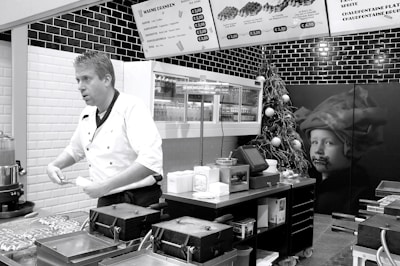 A chef in a white uniform is standing in a modern kitchen or food stall, possibly preparing food. Wall-mounted menu boards display food items and prices. The decor includes black and white tiles, and there is a Christmas tree with ornaments as well as a large picture of a child on the wall.