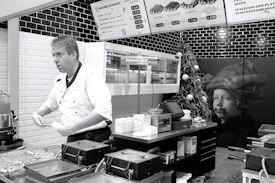 A chef in a white uniform is standing in a modern kitchen or food stall, possibly preparing food. Wall-mounted menu boards display food items and prices. The decor includes black and white tiles, and there is a Christmas tree with ornaments as well as a large picture of a child on the wall.