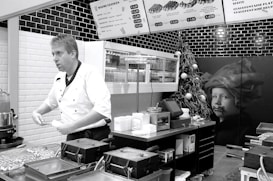 A chef in a white uniform is standing in a modern kitchen or food stall, possibly preparing food. Wall-mounted menu boards display food items and prices. The decor includes black and white tiles, and there is a Christmas tree with ornaments as well as a large picture of a child on the wall.
