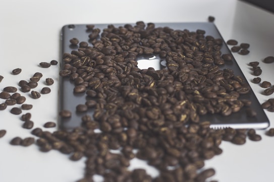 A large number of coffee beans are scattered over a sleek electronic device, possibly a tablet or laptop, with a visible apple logo.