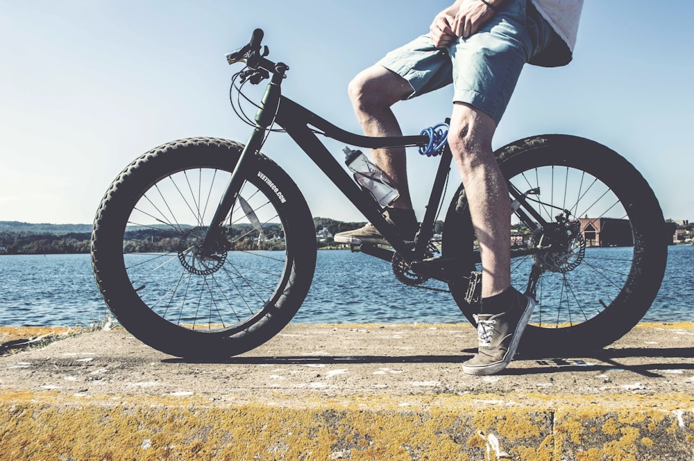 Homem na bicicleta de gordura preta na plataforma de concreto marrom com vista para o corpo de água durante o dia