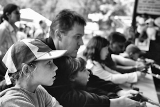 A group of people, including children and adults, are engaged in an activity at a fair or carnival. They appear focused and attentive, interacting with what seems to be a shooting game. The setting is outdoors, indicated by the blurred background suggesting a park-like environment.
